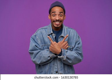 Overjoyed young brown-eyed dark skinned bearded male looking amazedly at camera with wide smile, showing in different sides with raised index fingers, isolated over purple background