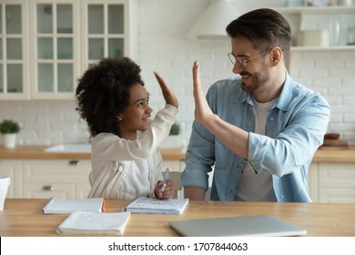 Overjoyed Young Caucasian Father And Ethnic Small Daughter Give High Five Finish Homework, Happy Dad And Biracial Little Girl Child Have Fun Study Together On Home Kitchen, Homeschooling Concept