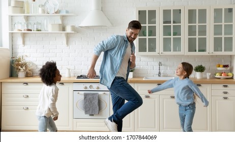 Overjoyed Young Caucasian Father And Little Multiracial Daughters Dancing Together In Home Kitchen, Happy Dad And Small Multiethnic Girls Children Have Fun Playing On Weekend Or Quarantine