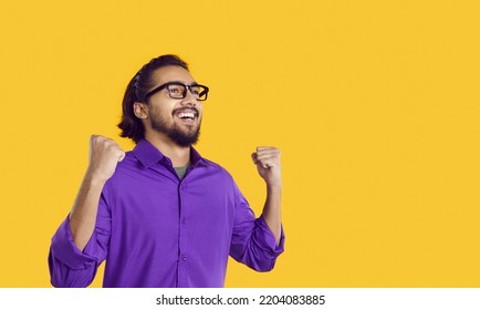 Overjoyed Young Dark Haired South Asian Man Raises Hands As Sign Victory Rejoice At Successful Events In Career Or Own Business And Looks Into Distance Smiling Standing In Yellow Studio. Copy Space