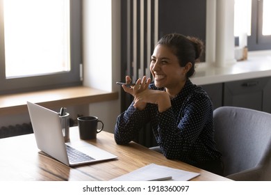 Overjoyed Young Ethnic Woman Sit At Home Office Work On Laptop Have Call On Cellphone Loudspeaker. Smiling Indian Female Talk Activate Smartphone Digital Voice Assistant. Modern Communication Concept.