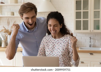 Overjoyed Young Family Couple Stand At Kitchen Look At Laptop Screen Celebrate Receiving Loan Approval From Bank. Excited Spouses Real Estate Agency Clients Get Email Offering House Of Dream For Sale