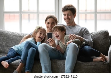 Overjoyed young family with small children sit relax on sofa at home laugh watching funny video on smartphone together, happy Caucasian parents rest with little kids using modern cellphone gadget