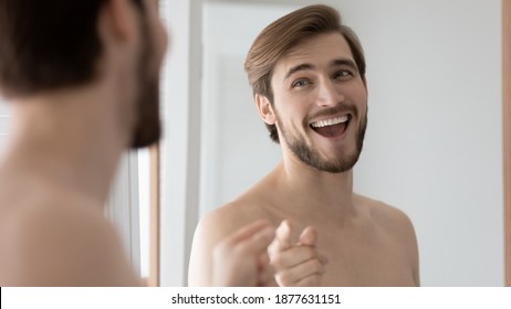 Overjoyed young handsome caucasian 30s man with healthy smile making gesture, looking in mirror, training self-confidence in morning after showering or setting positive mood in bathroom, head shot.