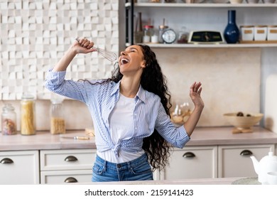 Overjoyed Young Lady Singing At Appliance As Imaginary Microphone, Dancing In Kitchen Interior And Enjoying Time Alone, Free Space. Fooling At Home Concept