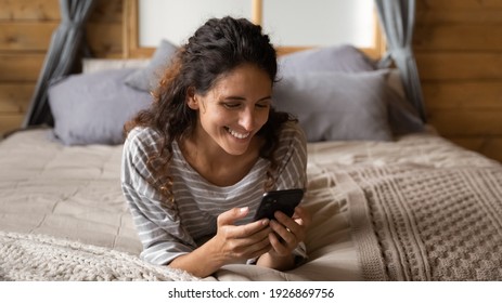 Overjoyed young Latino woman relax in bedroom use cellphone texting or messaging online. Happy millennial Hispanic female look at smartphone screen browse internet. Technology concept.