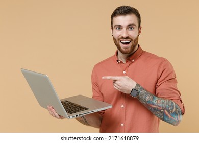 Overjoyed Young Man 20s He Wears Orange Shirt Hold In Hand Work On Laptop Pc Computer Pointing On It Look Camera Isolated On Plain Pastel Light Beige Background Studio Portrait. Tattoo Translate Fun