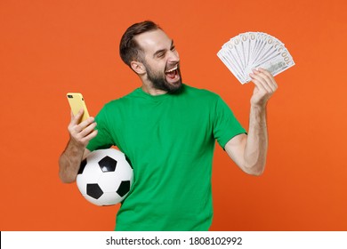 Overjoyed young man football fan in green t-shirt cheer up support favorite team with soccer ball using mobile cell phone fan of cash money isolated on orange background. People sport leisure concept