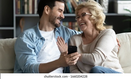 Overjoyed young man sit on couch laugh have fun using cellphone with mature mom, happy middle-aged 70s mother enjoy family weekend with adult son, watch funny video on smartphone together