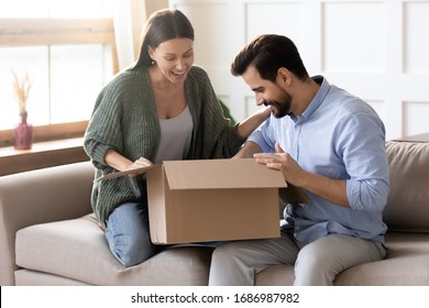 Overjoyed Young Married Couple Sitting On Sofa, Unpacking Cardboard Box At Home. Happy Family Spouses Clients Satisfied With Purchase In Internet Store Or Fast International Shipping Delivery.