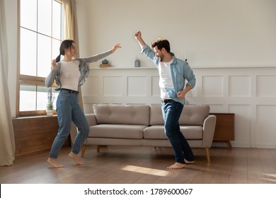 Overjoyed Young Married Family Couple Homeowners Dancing To Music Barefoot On Wooden Floor, Celebrating Moving Into New Apartment Or Freedom, Enjoying Active Domestic Hobby Pastime Together Indoors.