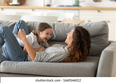 Overjoyed young mother lie on couch in kitchen have fun play with smiling little daughter enjoy weekend at home, happy mom feel playful engaged in childish activity with small preschooler girl child