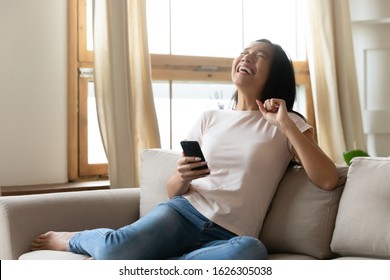 Overjoyed young Vietnamese woman sit relax on couch in living room laugh watch funny video on smartphone, happy millennial Asian girl rest on sofa at home have fun using modern cellphone device
