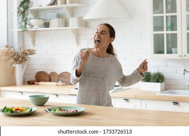 Overjoyed young woman cooking in modern design kitchen have fun dancing entertaining, happy smiling millennial girl preparing food in morning at home singing in appliance enjoy domestic weekend