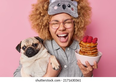 Overjoyed young woman with curly bushy hair exclaims loudly keeps mouth opened holds bowl of tasty pancakes with raspberry and pug dog awakes in good mood dressed in nightwear poses indoors.