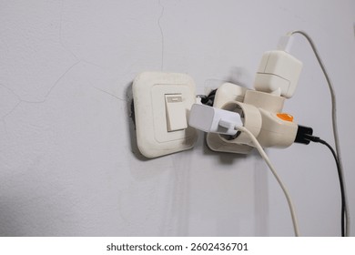 An overloaded electrical socket with multiple plugs and adapters connected to a power strip. The setup appears hazardous, with a tangled mess of cables and excessive power usage