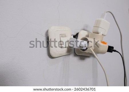 An overloaded electrical socket with multiple plugs and adapters connected to a power strip. The setup appears hazardous, with a tangled mess of cables and excessive power usage