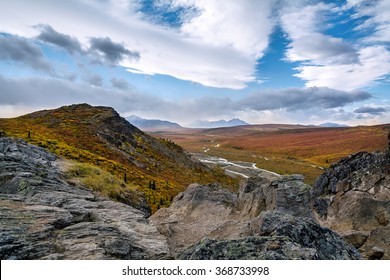 Overlooking Savage River in Denali National park during peak fall colors with oddly dramatic split in clouds; rocky foreground.
