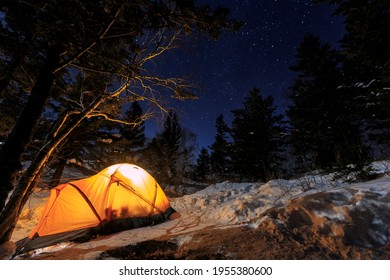 Overnight Stay At The Top Of The Winter Mountain. An Orange Tent, Illuminated From The Inside, Stands At The Edge Of The Forest In The Middle Of The Night In The Snow.