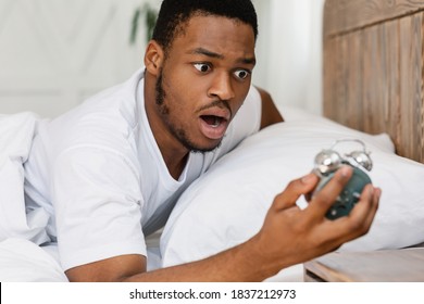 Oversleeping Concept. Shocked Black Guy Looking At Broken Alarm Clock Waking Up Late For Work In The Morning, Lying In Bed At Home. Awakening Problem and Tardiness, Bad Morning