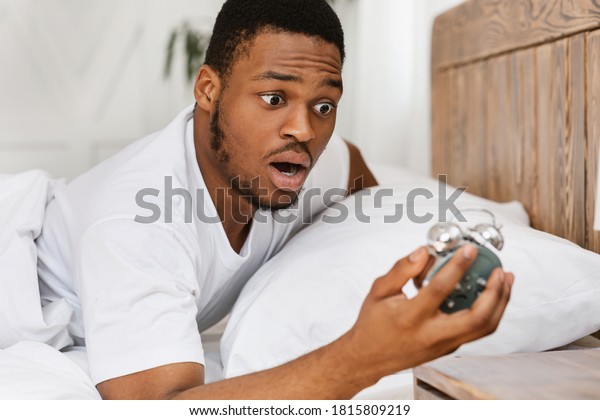 Oversleeping.\
Overslept African American Man Looking In Shock At Broken Alarm\
Clock Waking Up In The Morning, Lying In Bed At Home. Late For\
Work, Bad Morning, Tardiness\
Concept