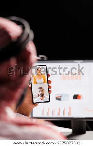 Over-the-shoulder image of an Arab guy using a digital mobile gadget to talk to others. Detailed shot of a Muslim man using his smartphone to conduct a video conference call while seated at table.