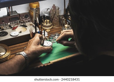 An over-the-shoulder view of a watchmaker repairing the machinery of a watch on his workbench full of various specialised tools of the watchmaking trade. - Powered by Shutterstock