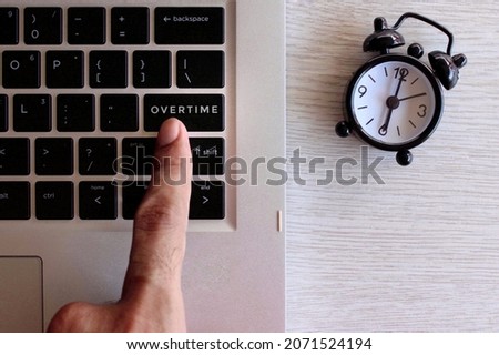 Overtime work concept. Finger pressing keyboard with text OVERTIME and black alarm clock.