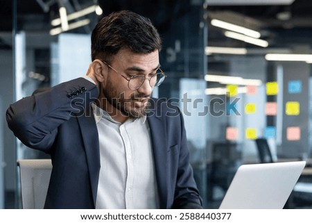 Overtired and overworked man at workplace inside office in business suit. Businessman suffering from severe neck pain.