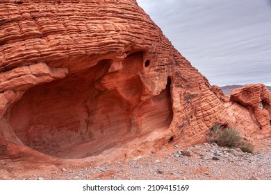 Overton, Nevada, USA - February 24, 2010: Valley Of Fire. Closeup Of Big Hole Or Cave In Lined And Cracked Red Rock On Dry Desert Floor Under Thick Gray Sky.