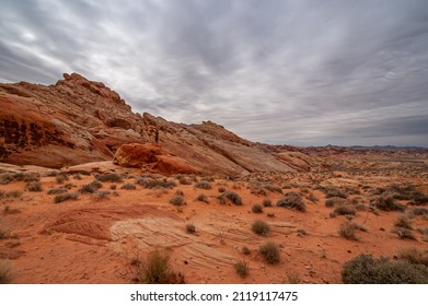 Overton, Nevada, USA - February 25, 2010: Valley Of Fire. Wide Landscape With Slanted Mountain Outcrop Raising Out Of Dry Red Rock Desert Floor With Shrubs Under Heavy Gray Cloudscape.