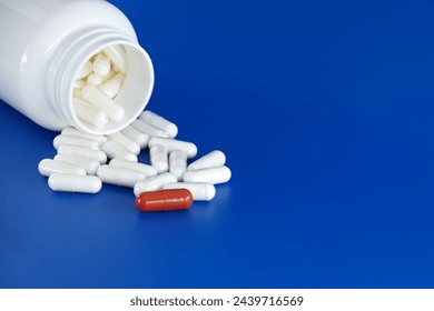 Overturned white medicine bottle with its contents white pills or capsules, spilled out onto a blue background. Among these white pills, a solitary red pill stands out