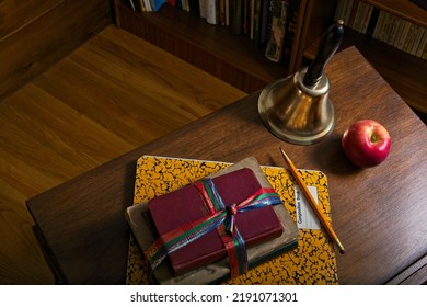 Overview Of Antique Wooden School Desk Concept With Bookstrap Wrapped Textbooks, A Yellow Composition Book, A Red Apple And Brass Hand Bell. The Desk Is Side Lit, Stands Beside Wood Bookcases