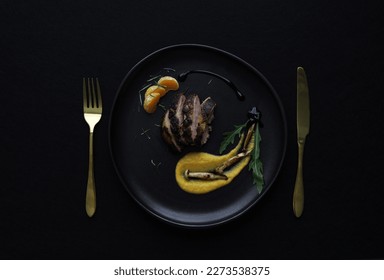 Overview of a black plate in a black background with roasted duck breast, squash purée, mushrooms and orange. Golden fork and knife. Dark mood photo in professional studio lighting of a fine dining.