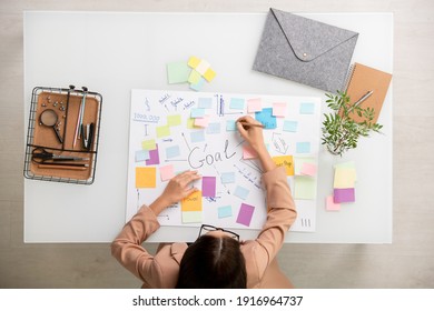 Overview of brunette businesswoman making working notes while bending over large document with main goals and points on sticky notepapers