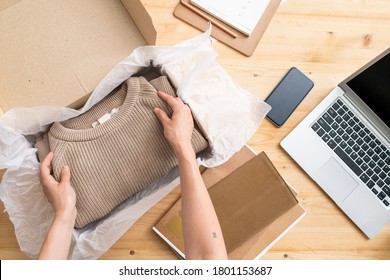 Overview of hands of young female manager of online shop putting folded beige knitted sweater into box before packing it and sending to client