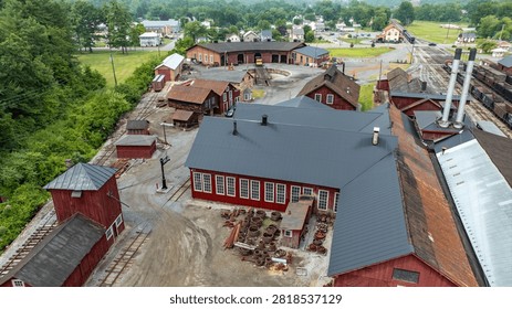 An overview of a historical train yard with multiple red workshop buildings and a simple landscape. The area is surrounded by trees and some small town buildings are nearby.