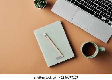 Overview of laptop keyboard, cup of coffee, notebook with pen and green plant in small flowerpot on workplace of modern business person