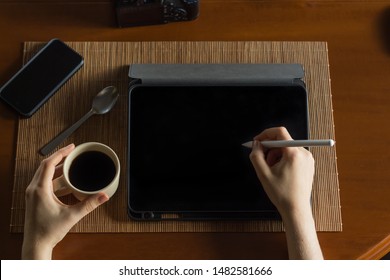 Overview of a woman working in a wooden desk, with a tablet and a stylus, having a coffee with other stuff