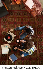 Overview Of Young Intercultural Couple In Stylish Casualwear Sitting On Red Carpet In Living Room And Listening Vynil Disks On Record Player