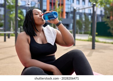 Overweight African Black Woman Sitting On Fitness Mat Having Rest Outdoors, Drinking Fresh Water, Enjoying Sport. Tired And Exhausted Female In Black Tracksuit Need Some Rest. Copy Space
