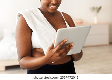 Overweight black lady with tablet pc watching sports video tutorial, checking music for domestic workout at home. Plus size African American woman with touch pad talking to personal trainer online - Powered by Shutterstock