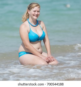 Overweight blonde at the sea. Adult woman in bikini sitting in waters edge with hands on knees and looking at camera smiling, square composition