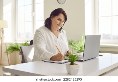 Overweight brunette young woman using laptop in modern office. Focused plus size woman reviewing financial documents and making notes on paper. Financial manager, bookkeeper, accountant - Powered by Shutterstock