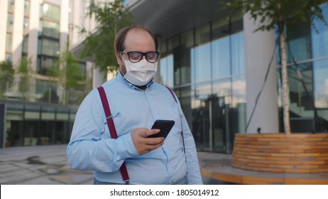 Overweight Businessman On Medical Mask Using Smartphone Walking Near Office Building. Obese Employee Wearing Protective Mask Texting Email On Smartphone Walking Outdoors