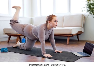 Overweight Caucasian Woman Doing Sport Exercises On Fitness Mat At Home, Using Laptop. Fat Redhead Lady In Sportive Outfit Raising One Leg Up. Fitness, Home Training, Technology And Diet.