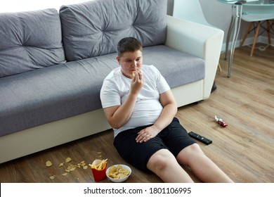 An Overweight Child Boy Enjoy Leading Unhealthy Passive Lifestyle, Sits On The Floor Watching Tv And Eating, He Moves A Little That Is Why He Is Fat