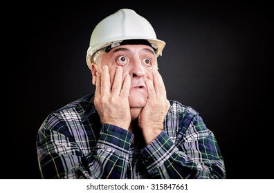 Overweight constructor worker with eyes wide open, hopeless gestures. Hands on face, all over black background.