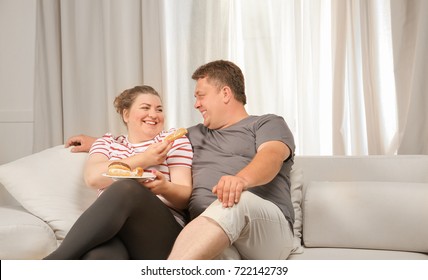 Overweight Couple Eating Sweets On Sofa At Home