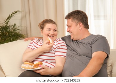 Overweight Couple Eating Sweets On Sofa At Home